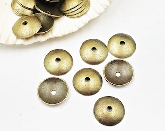 Può includere: Un gruppo di 10 perline distanziatrici in metallo color oro antico con una parte superiore a cupola e un piccolo foro al centro. Le perline sono sparse su una superficie bianca.