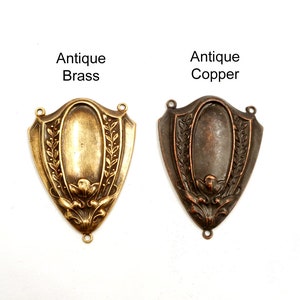 Puede incluir: Dos colgantes antiguos de metal con forma de escudo, uno de lat&oacute;n y otro de cobre, con dise&ntilde;os florales y de hojas. El colgante de lat&oacute;n est&aacute; etiquetado como "Antique Brass" y el de cobre como "Antique Copper". Ambos tienen bucles para sujetar.