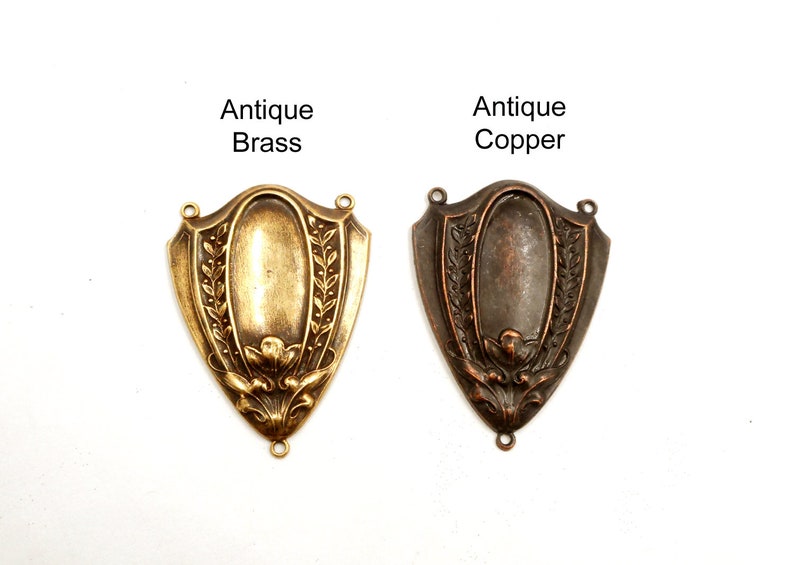 Puede incluir: Dos colgantes antiguos de metal con forma de escudo, uno de lat&oacute;n y otro de cobre, con dise&ntilde;os florales y de hojas. El colgante de lat&oacute;n est&aacute; etiquetado como "Antique Brass" y el de cobre como "Antique Copper". Ambos tienen bucles para sujetar.