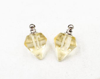 May include: Two diamond-shaped perfume bottles with silver-colored stoppers. The bottles are clear with a yellow tint, and the stoppers have a small, round design. The bottles are displayed on a white surface.