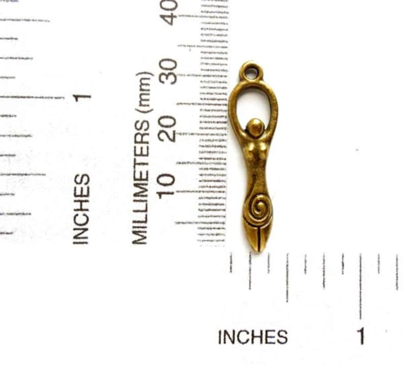 May include: A gold-toned metal charm depicting a stylized female figure with a spiral design. The charm is approximately 1 inch in length and is shown next to a ruler for size reference.