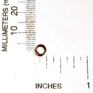 May include: A small, copper-colored metal ring, approximately 0.25 inches in diameter, sits on a white surface. The ring is next to a ruler marked in inches and millimeters, providing scale. The ring appears to be a component or finding.