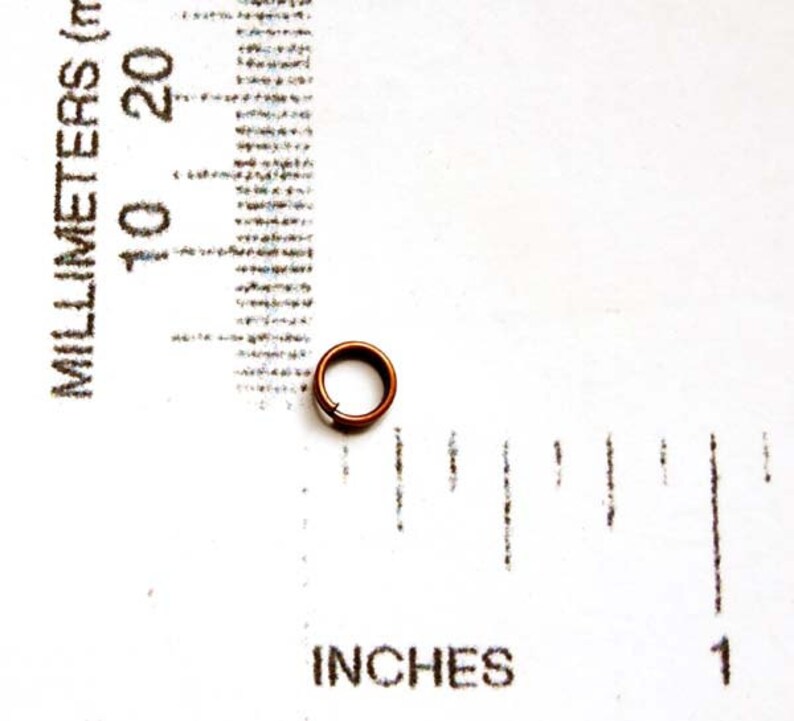 May include: A small, copper-colored metal ring, approximately 0.25 inches in diameter, sits on a white surface. The ring is next to a ruler marked in inches and millimeters, providing scale. The ring appears to be a component or finding.