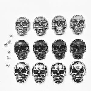 May include: An overhead shot shows twelve skull-shaped metal embellishments in varying shades of silver and black. The skulls are arranged in three rows of four, with several small, round silver rivets to the left. The skulls have detailed facial features.