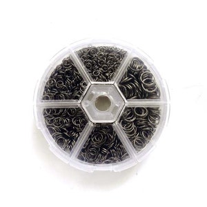 May include: A clear plastic container with six compartments filled with small silver jump rings. The container is round and has a hexagonal center.