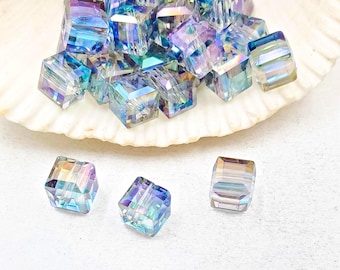 May include: Iridescent cube-shaped beads in shades of blue, purple, and gold. Some beads are scattered on a white seashell, while others are on a white surface. These beads are ideal for jewelry making and crafting.