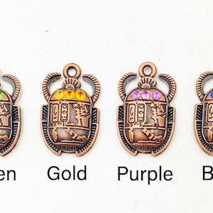 May include: Four bronze-colored scarab beetle charms with colorful accents. Each charm features a different color of gemstone: green, gold, purple, and blue. The charms have an Egyptian theme and are labeled with their respective colors.