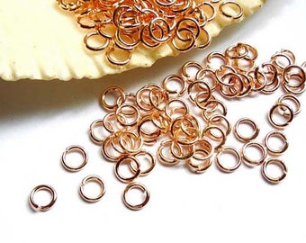 May include: A large pile of gold-toned jump rings, a type of jewelry finding, are scattered on a white surface. The jump rings are small and round, and they are perfect for connecting different pieces of jewelry together.