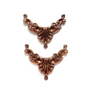 May include: Two ornate copper-colored metal embellishments with a decorative, floral design. Each piece has three holes for attaching to a project.