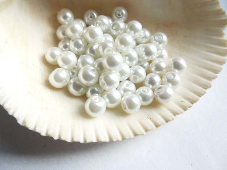 May include: A pile of white pearl beads in a seashell. The beads are round and smooth, and they are arranged in a random pattern.