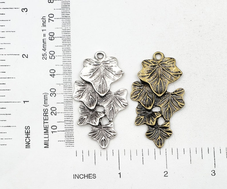 May include: Two metal charms in the shape of ivy leaves. The charm on the left is silver and the charm on the right is gold. Both charms are approximately 2 inches long.