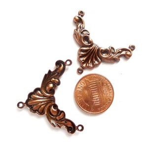 May include: Two antique copper-colored metal corner embellishments with a decorative floral design. The embellishments are approximately the size of a US penny.