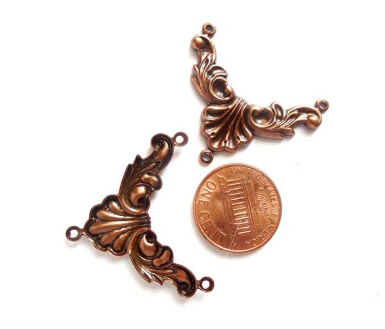 May include: Two antique copper-colored metal corner embellishments with a decorative floral design. The embellishments are approximately the size of a US penny.