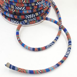 May include: A spool of colorful patterned cord with a blue, red, white, and green geometric design. The cord is wound around a cardboard spool and a length of the cord is draped over the spool.