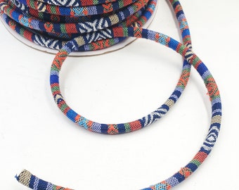 May include: A spool of colorful patterned cord with a blue, red, white, and green geometric design. The cord is wound around a cardboard spool and a length of the cord is draped over the spool.