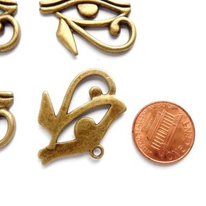 May include: Five antique gold-toned metal charms in the shape of the Eye of Horus, an ancient Egyptian symbol. The charms are approximately the size of a US penny.