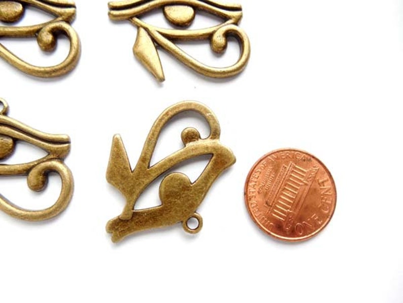 May include: Five antique gold-toned metal charms in the shape of the Eye of Horus, an ancient Egyptian symbol. The charms are approximately the size of a US penny.