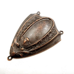 Puede incluir: Un componente met&aacute;lico decorativo en forma de escudo con un centro ovalado y detalles florales y de hojas ornamentados. La pieza tiene un acabado en bronce oscuro y peque&ntilde;os bucles para la fijaci&oacute;n.