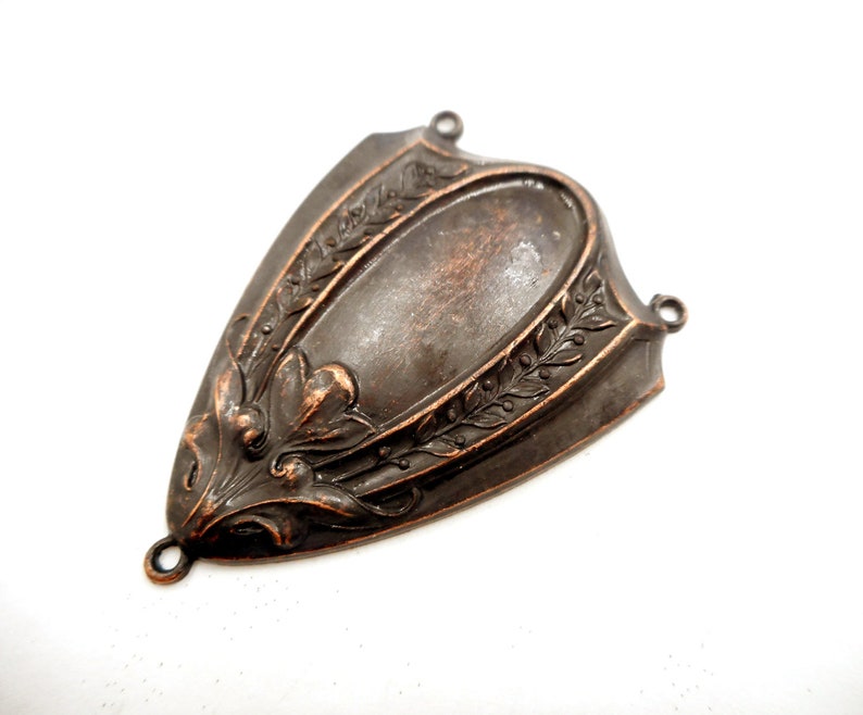 Puede incluir: Un componente met&aacute;lico decorativo en forma de escudo con un centro ovalado y detalles florales y de hojas ornamentados. La pieza tiene un acabado en bronce oscuro y peque&ntilde;os bucles para la fijaci&oacute;n.