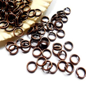 May include: Close-up of a collection of copper-colored jump rings scattered on a white surface. The rings are small, circular, and have a split in the metal. Some rings are piled on a seashell.