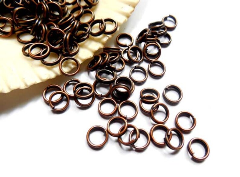 May include: Close-up of a collection of copper-colored jump rings scattered on a white surface. The rings are small, circular, and have a split in the metal. Some rings are piled on a seashell.