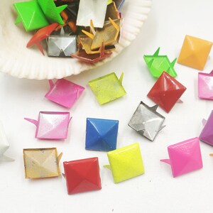 May include: Assortment of colorful square pyramid shaped metal studs with a pointed base. The studs are in various colors including pink, green, blue, yellow, red, silver, and gold.