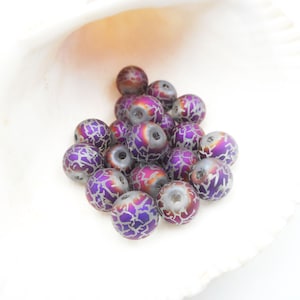 May include: A collection of round beads with a metallic finish. The beads have a base color of gray with vibrant purple and orange hues. Each bead is adorned with a silver, crackle-like pattern, creating a unique texture. The beads are displayed against a white background.