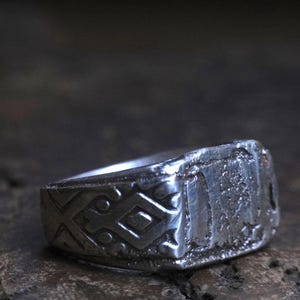 May include: A silver-colored ring with a rectangular face and geometric patterns. The ring has a worn, antique appearance, suggesting it may be vintage or handcrafted. The design includes a series of intersecting lines and shapes.