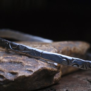 May include: A silver-colored bangle bracelet with an engraved floral pattern. The bracelet is resting on a textured, brown stone surface, with a dark background. The bracelet's design features repeating curved lines and shapes.