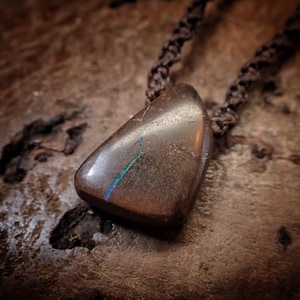 May include: A brown pendant necklace featuring a polished, triangular stone with a blue and green streak. The stone hangs from a braided brown cord. The background is a textured, dark brown surface.