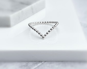 Puede incluir: Un anillo plateado en forma de V, compuesto por pequeñas esferas espaciadas uniformemente. El anillo se encuentra sobre una superficie blanca, con una estructura similar al mármol en el fondo. El diseño del anillo es simple y elegante, adecuado para el uso diario.