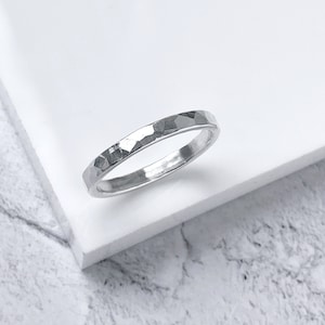 May include: A silver ring with a hammered texture. The ring is thin and has a faceted surface, reflecting light. The ring is displayed on a white surface.