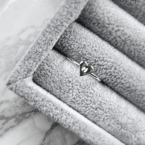 Könnte beinhalten: Ein silberner Ring mit einem herzförmigen Anhänger, der den Buchstaben "M" in der Mitte zeigt. Der Ring wird in einer grauen Samt-Schmuckschatulle präsentiert. Der Hintergrund ist eine Marmoroberfläche.