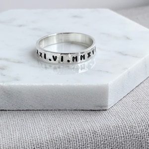 May include: A silver ring with Roman numerals engraved around the band. The ring has a hammered texture and sits on a white marble surface. The ring is a simple, elegant piece of jewelry.