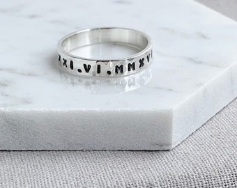 May include: A silver ring with Roman numerals engraved around the band. The ring has a hammered texture and sits on a white marble surface. The ring is a simple, elegant piece of jewelry.