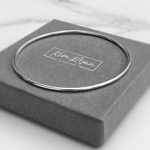 May include: A silver bangle bracelet on a grey jewellery box. The bracelet is a thin, polished band. The box features the text "Kim Ryan Jewellery" in a white frame. The overall aesthetic is minimalist.