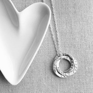 Könnte beinhalten: Eine silberne Halskette mit drei ineinandergreifenden Ringen, die jeweils mit Worten graviert sind. Die Ringe hängen an einer filigranen Kette. Eine weiße herzförmige Schale befindet sich im Hintergrund auf einer strukturierten Oberfläche.