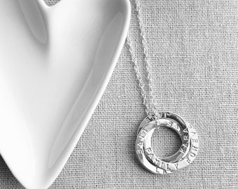May include: A silver necklace with three interlocking rings, each engraved with words. The rings hang from a delicate chain. A white heart-shaped dish is in the background, resting on a textured surface.