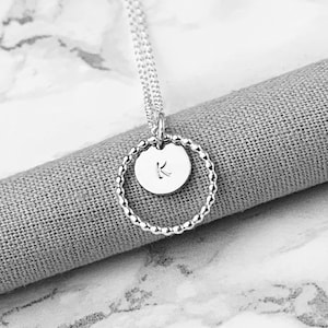 Op de afbeelding: Een zilveren ketting met een hanger in de vorm van een cirkel met kralen en een kleinere schijfhanger. De schijfhanger heeft de letter "K". De ketting wordt getoond op een grijze stoffen ondergrond.