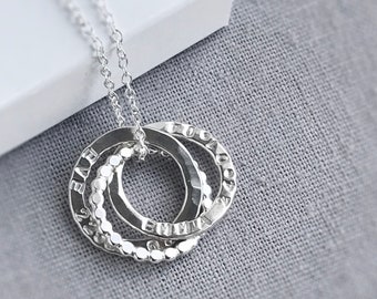 May include: A silver necklace featuring three interlocking rings. Each ring has a different texture and is engraved with a name. The necklace hangs on a delicate chain, displayed on a gray fabric background.