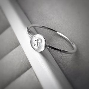 May include: A silver ring with a small, round, engraved charm. The charm features a stylized letter "J". The ring band is thin and polished, and the charm is centered on the band. The ring is set against a soft grey background.