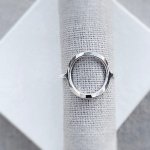 Op de afbeelding: Een zilveren ring met een open ovaal ontwerp. De ring heeft een getextureerde, gehamerde uitstraling. De ring wordt getoond op een grijze stof, wat de minimalistische en moderne esthetiek benadrukt. De ring is een sieraad.