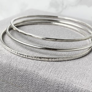 May include: Four silver bangles with different surface finishes. One has a hammered effect, another a textured pattern, and the others are smooth. The bangles are presented on a grey fabric.