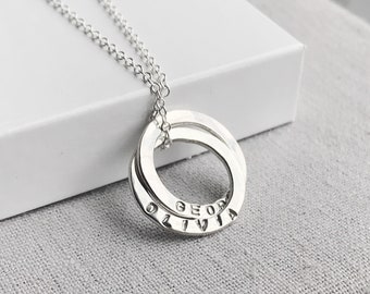 May include: A silver necklace featuring two interlocking rings. One ring is engraved with the name "OLIVIA", and the other with "GEORGIA". The necklace hangs on a delicate silver chain, displayed against a white box and a gray fabric background.