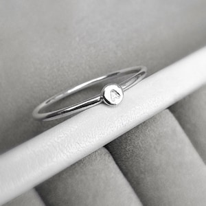 Op de afbeelding: Een fijne zilveren ring met een kleine, ronde bedel met de letter "A". De ring heeft een minimalistisch ontwerp, geschikt voor dagelijks gebruik. De ring wordt getoond op een wit oppervlak tegen een zachte grijze achtergrond.