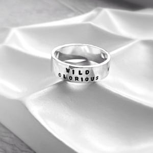 May include: A silver ring with the words "WILD GLORIOUS" stamped on the outside. The ring is set against a textured white surface, creating a contrast between the shiny metal and the matte background. The ring is a simple band design.