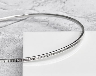 May include: A silver-colored bangle bracelet with a textured, hammered design. The bracelet is round and appears to be made of metal. The textured portion of the bracelet is approximately 0.25 inches wide. The background is white and gray.