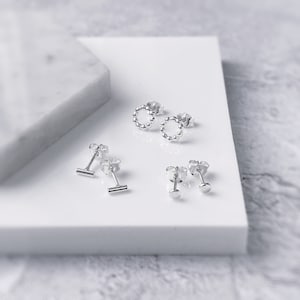 Könnte beinhalten: Ein Set mit vier silbernen Ohrsteckern. Zwei Ohrstecker sind kleine Kreise mit einem perlenbesetzten Rand, ein Ohrstecker ist eine kleine Stange und der andere Ohrstecker ist eine kleine runde Scheibe.