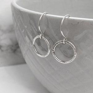 Peut inclure: Une paire de boucles d'oreilles créoles en argent avec des détails texturés. Les boucles d'oreilles sont circulaires avec un design simple en forme de crochet. Les boucles d'oreilles sont présentées sur un fond gris clair.