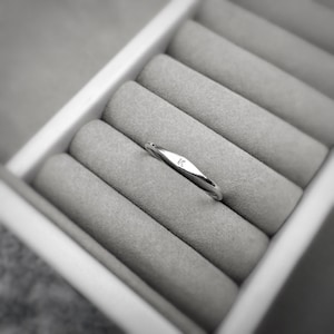 May include: A silver ring with a small, elongated design, featuring the letter "K" engraved on its surface. The ring is set against a gray, velvet-lined jewelry box, creating a minimalist aesthetic. The ring is a delicate piece of jewelry.